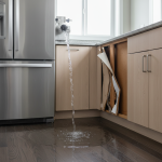 A close-up view of severe water damage impacting the base of kitchen cabinets, visible as swollen wood and discolored laminate. This common damage often results from a hidden ice maker filter leak, originating from a faulty refrigerator water filter manifold, a frequent cause of catastrophic leaks from aftermarket filters.